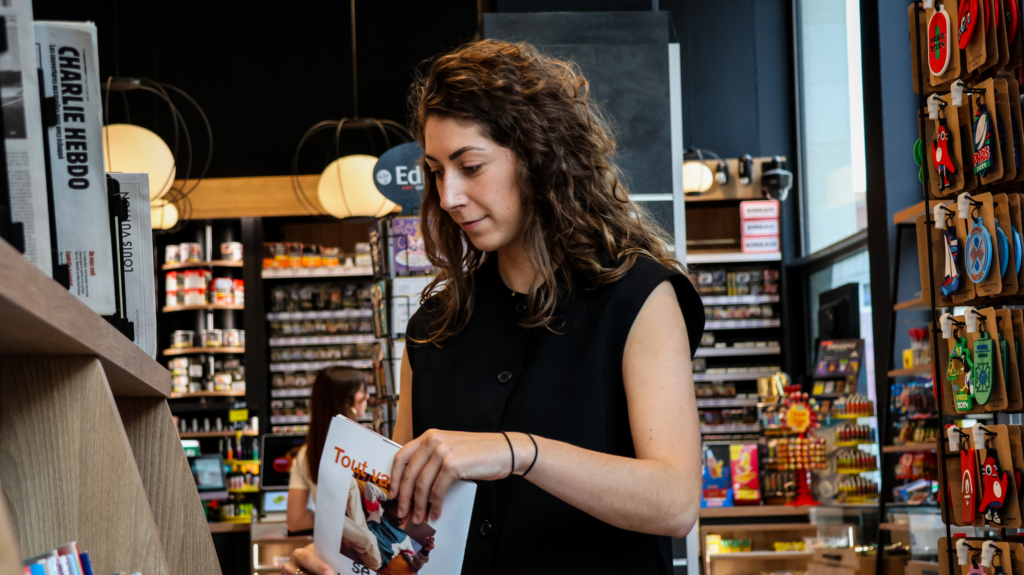 jeune femme au rayon presse d'un commerce buraliste. Elle tient un magazine dans les mains et permet d'illustrer le métier de vendeur commercial parcours buraliste